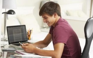 Teenage Boy Studying At Desk In Bedroom Using Mobile Phone